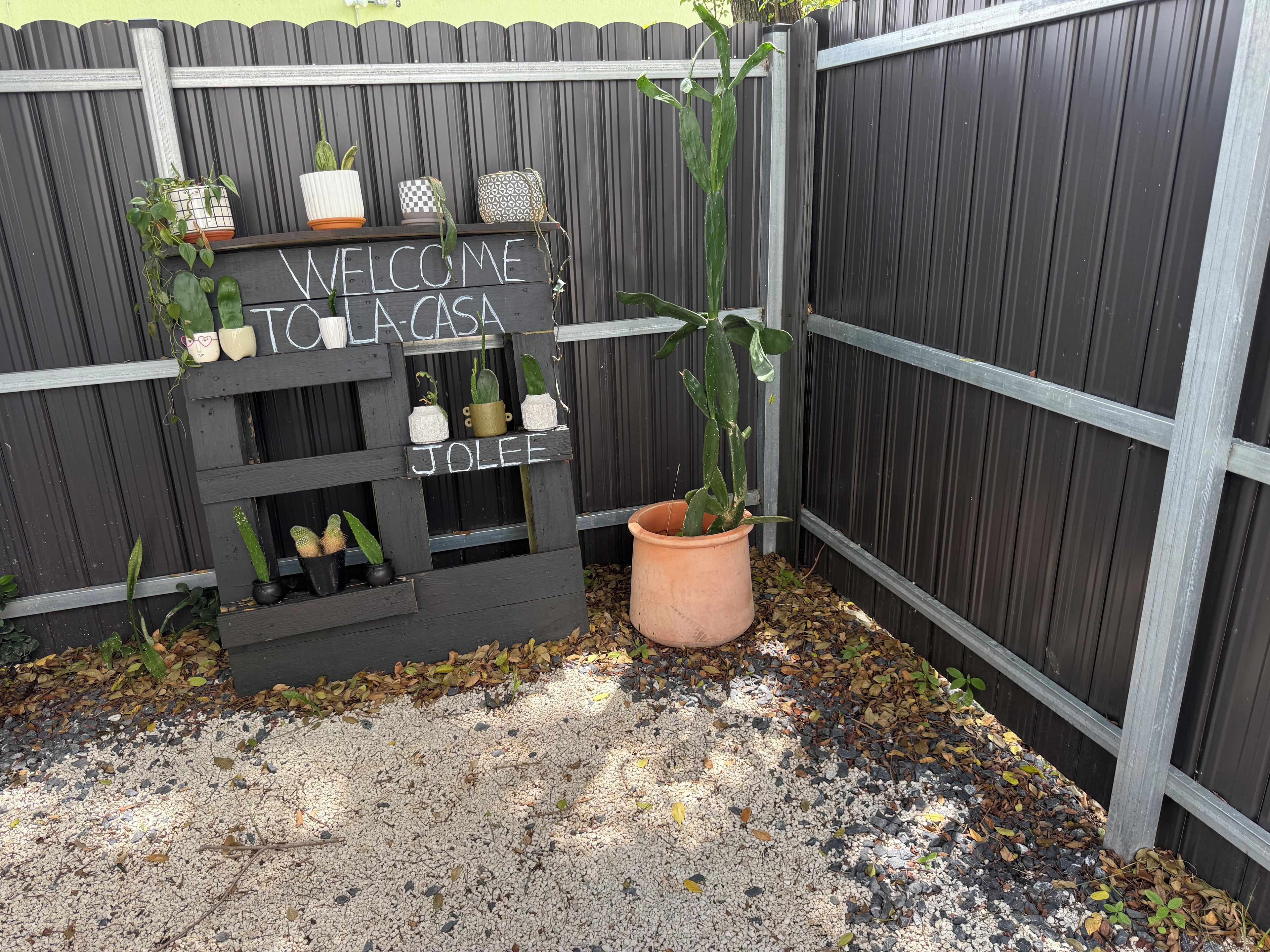Uma placa de madeira com a inscrição 'WELCOME TO LA CASA' e 'JOLEE', rodeada por plantas em vasos.