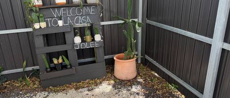 Uma placa de madeira com a inscrição 'WELCOME TO LA CASA' e 'JOLEE', rodeada por plantas em vasos.