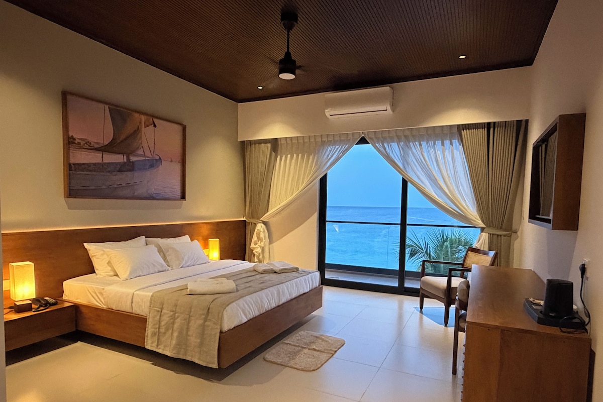 Habitación de hotel con una cama grande, lámparas de noche, un escritorio y vistas al mar a través de las ventanas.