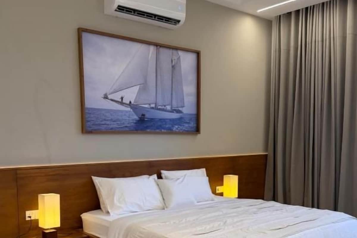 Habitación de hotel con cama, mesitas de noche, un cuadro de un velero, un equipo de aire acondicionado y una ventana con cortinas.