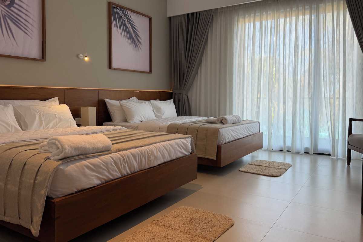 Una habitación de hotel con dos camas, un ventanal con cortinas, aire acondicionado de pared y un cuadro enmarcado.