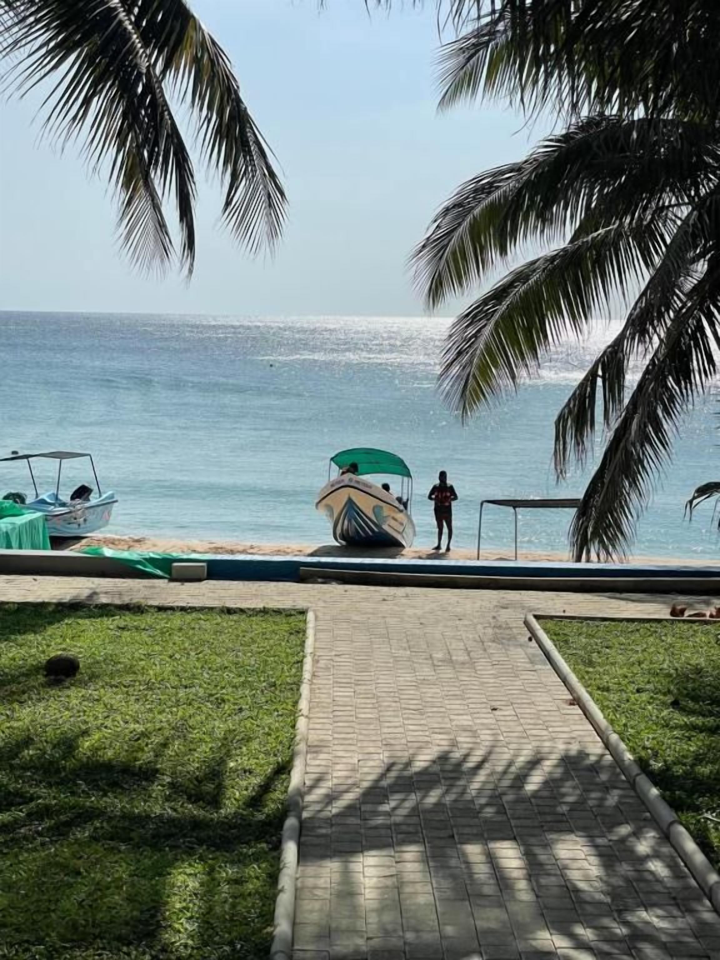 Un chemin pavé mène à une plage où se trouvent un bateau et une personne debout près de l’eau.