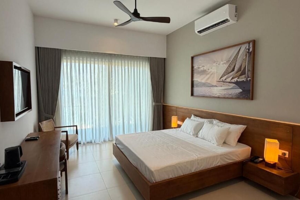 A hotel room with a bed, bedside lamps, a desk with a chair, a ceiling fan, and a large window with curtains.