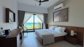 A hotel room with a bed, a desk, a chair, a ceiling fan, an air conditioning unit, and a large window with a view of the sea.