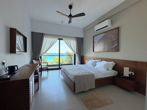 Habitación de hotel con cama, escritorio, silla, ventilador de techo, aire acondicionado y una ventana grande con vista al mar.