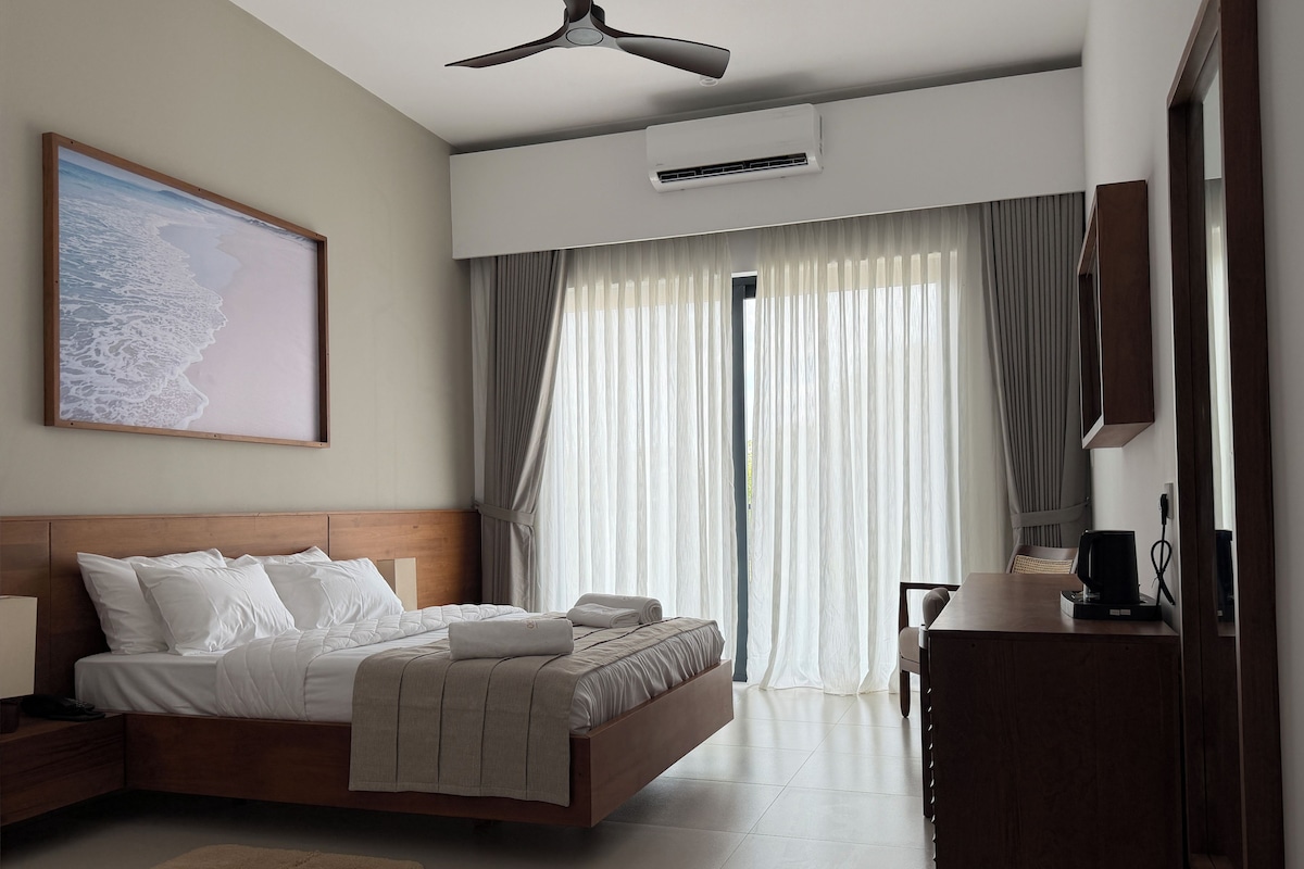 Una habitación de hotel con una cama, un escritorio, una silla, un ventilador de techo, un equipo de aire acondicionado y un amplio ventanal con cortinas.
