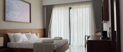 A hotel room with a bed, a desk, a chair, a ceiling fan, an air conditioning unit, and a large window with curtains.