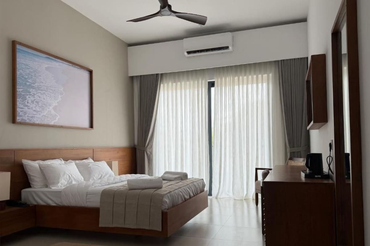 Un dormitorio moderno con cama, ventilador de techo, equipo de aire acondicionado, escritorio con silla y una ventana grande con cortinas.