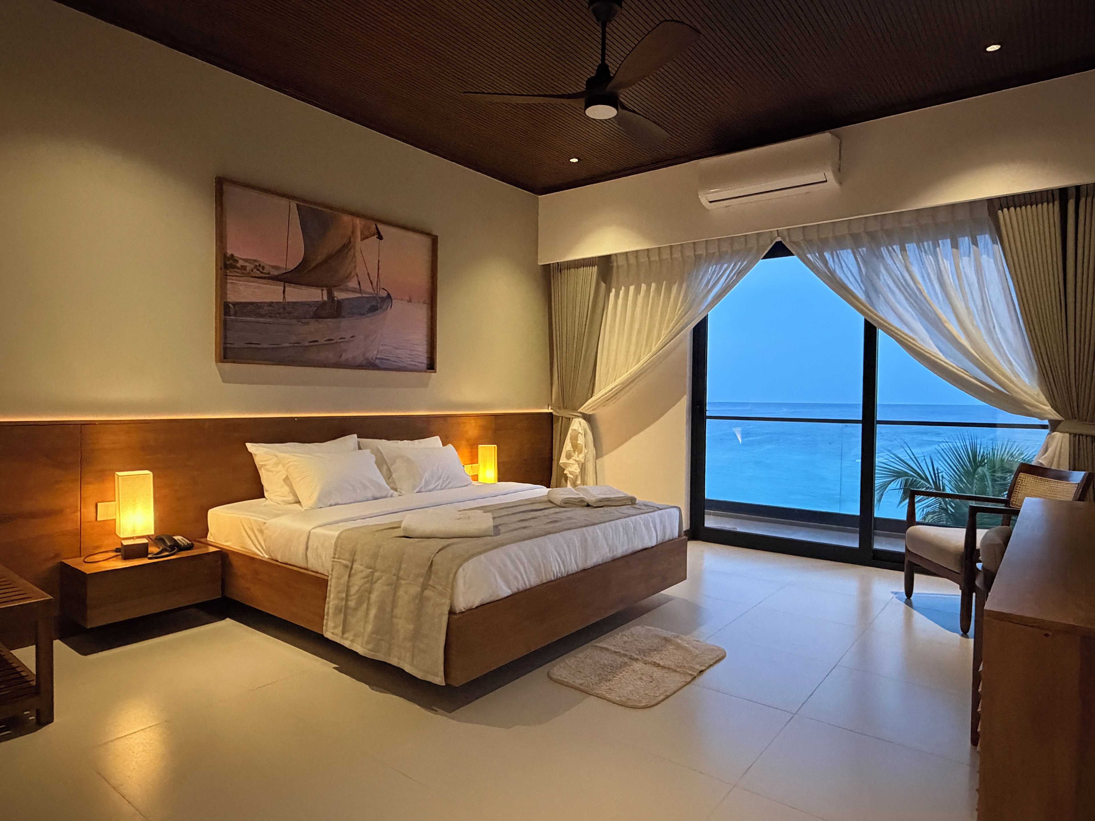 A bedroom with a bed, bedside lamps, a ceiling fan, a large window with curtains, and a painting on the wall.