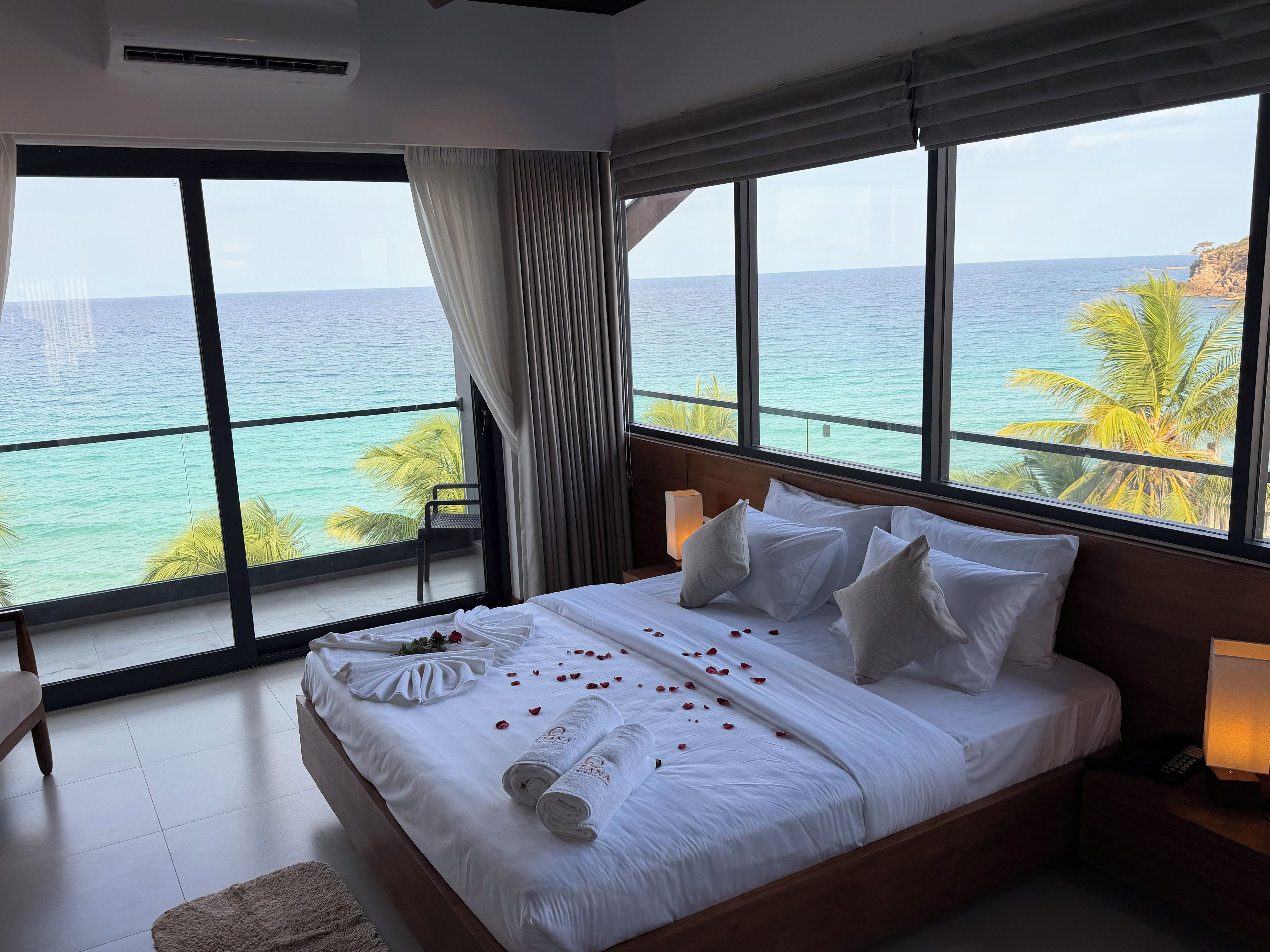 A hotel room with a large bed, a view of the sea, and a balcony with a chair.