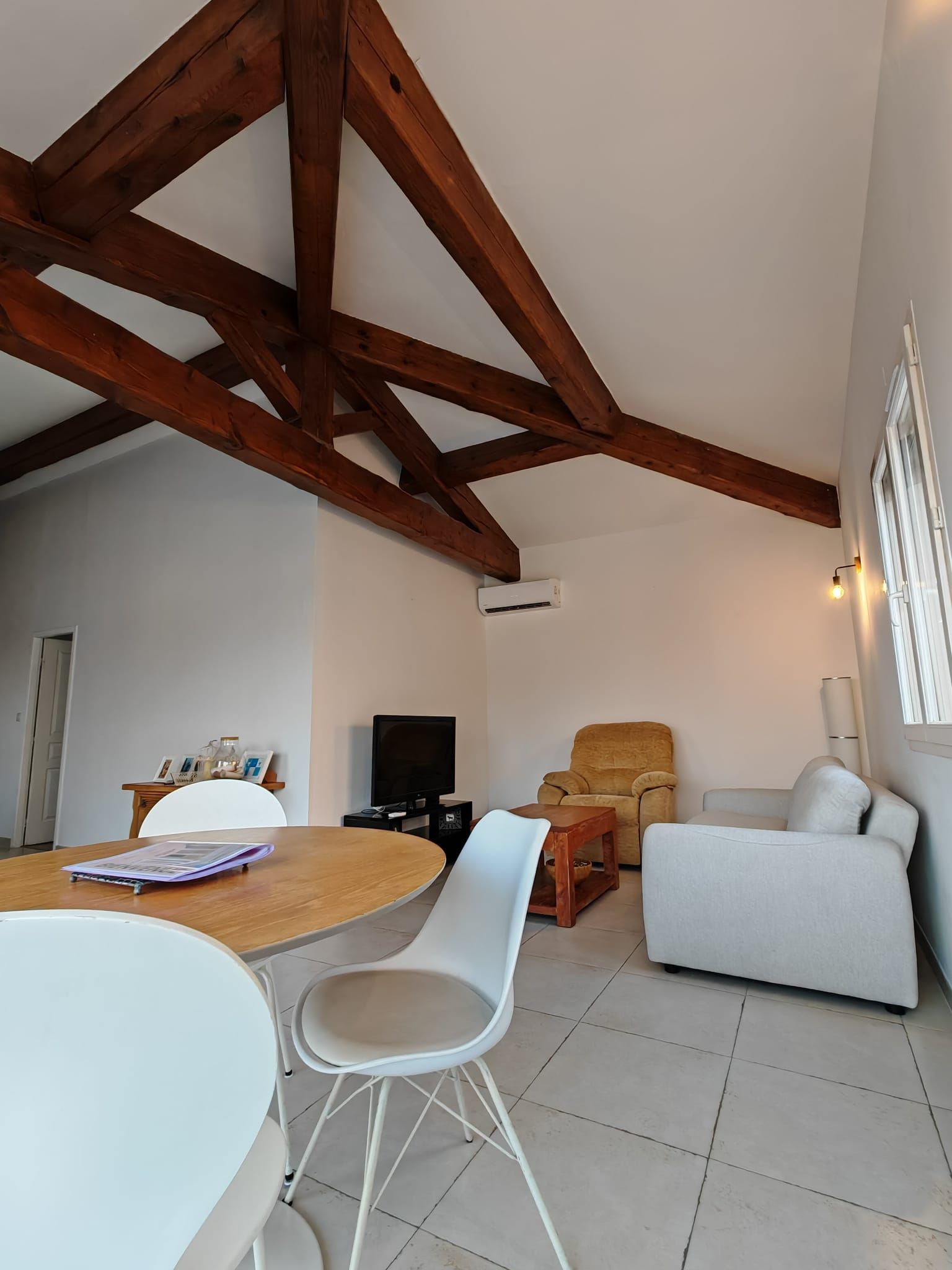 Un vaste espace de vie doté d’un plafond haut, de poutres en bois, d’un téléviseur, d’un canapé, d’un fauteuil et d’une table à manger.
