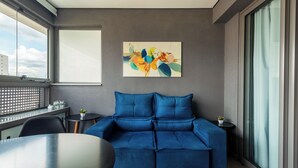 A modern living room with a blue sofa, a painting on the wall, and a view of the city through large windows. - Oscar by Bhomy (São Paulo)