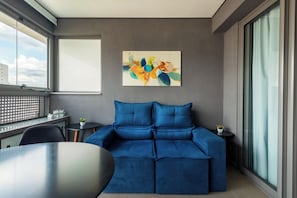 Ein modernes Wohnzimmer mit einem blauen Sofa, einem Gemälde an der Wand und Blick auf die Stadt durch große Fenster.