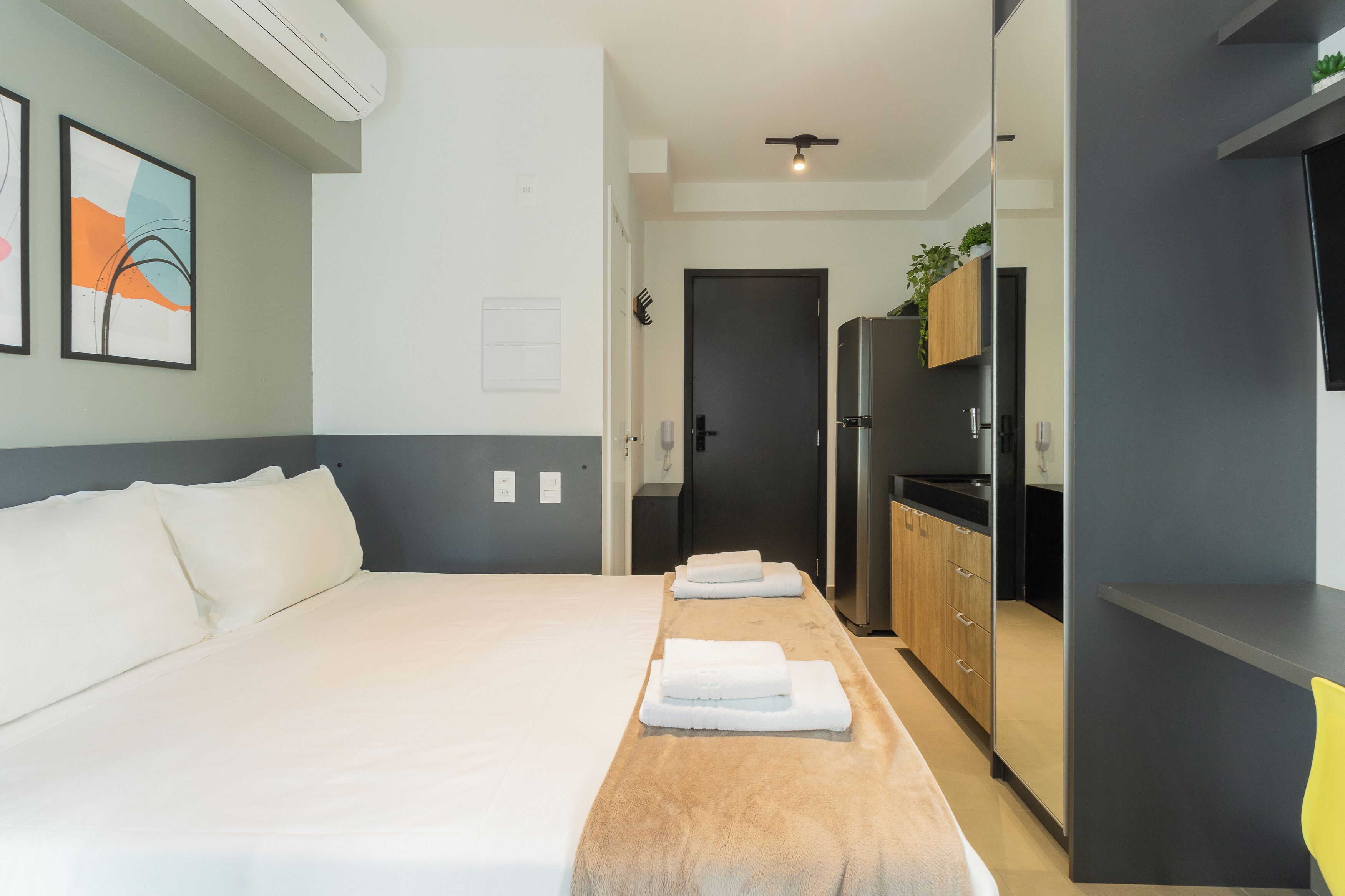 Une chambre d’hôtel comprenant un lit, une table de chevet en bois avec des serviettes, une kitchenette et une porte donnant sur une autre pièce.