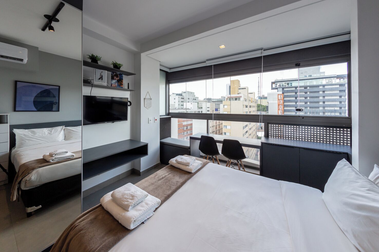 Habitación de hotel moderna con una cama grande, televisor montado en la pared, zona de estar con sillas y vista a la ciudad a través de la ventana.
