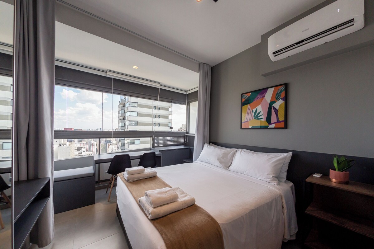 Ein Hotelzimmer mit einem großen Bett, einem Schreibtisch, einem Sessel und einem Fenster mit Blick auf Gebäude, sowie einer Klimaanlage.