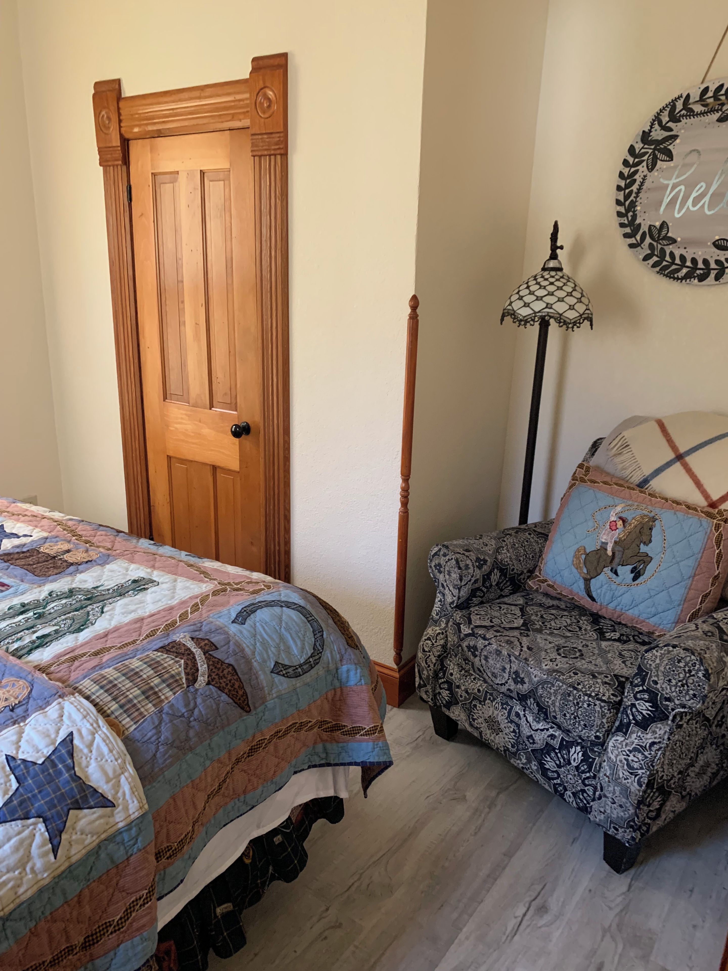 A bedroom with a wooden door, a patterned armchair, a quilted bedspread, and a wall-mounted lamp.