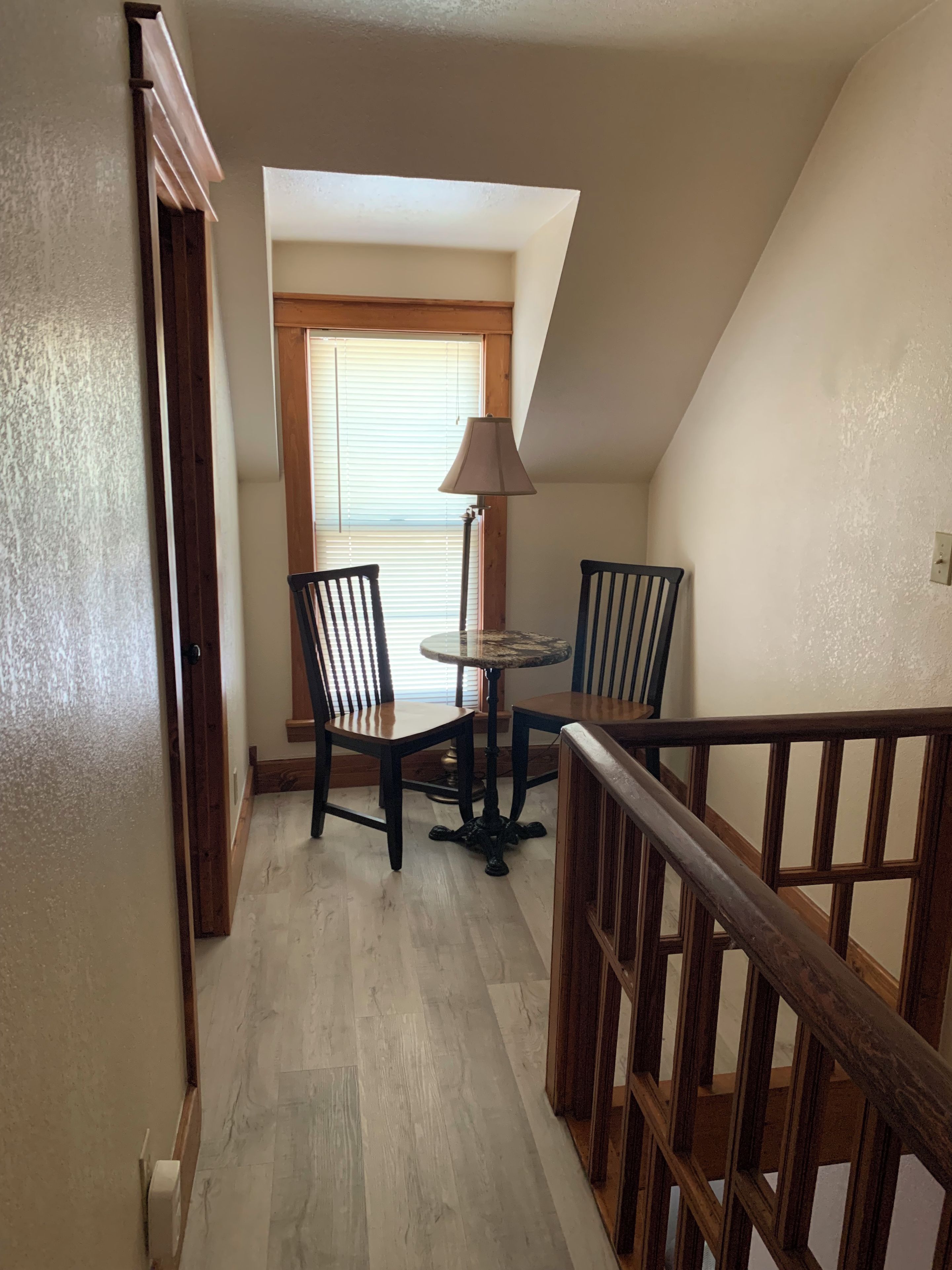 A staircase with wooden railings leading to a room with a table and chairs, a lamp, and a window with blinds.