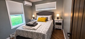 A bedroom with a bed, two bedside tables, a nightstand, a window with blinds, and an air conditioning unit. - The Northwood Nook on the Lake (Emily)