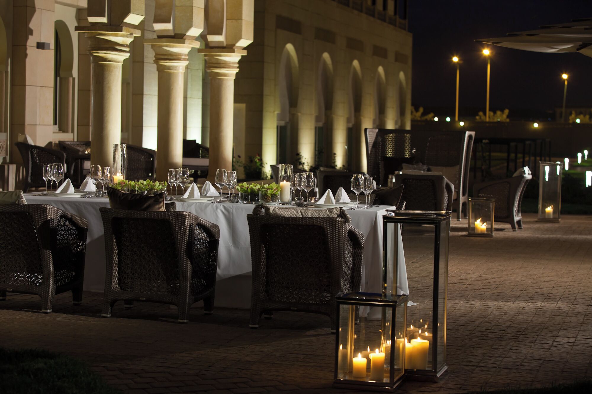 An outdoor dining area with tables set for a meal, featuring wicker chairs and candlelit centerpieces.