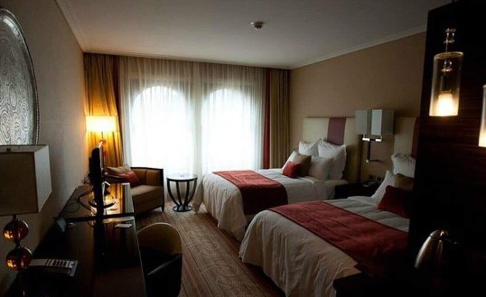 Une chambre d’hôtel avec deux lits, un bureau avec un ordinateur, une chaise, une lampe et une grande fenêtre avec des rideaux.