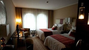 A hotel room with two beds, a desk with a computer, a chair, a lamp, and a large window with curtains.