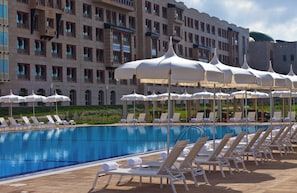 Una piscina con sillones y sombrillas, rodeada por un edificio con múltiples ventanas y balcones.