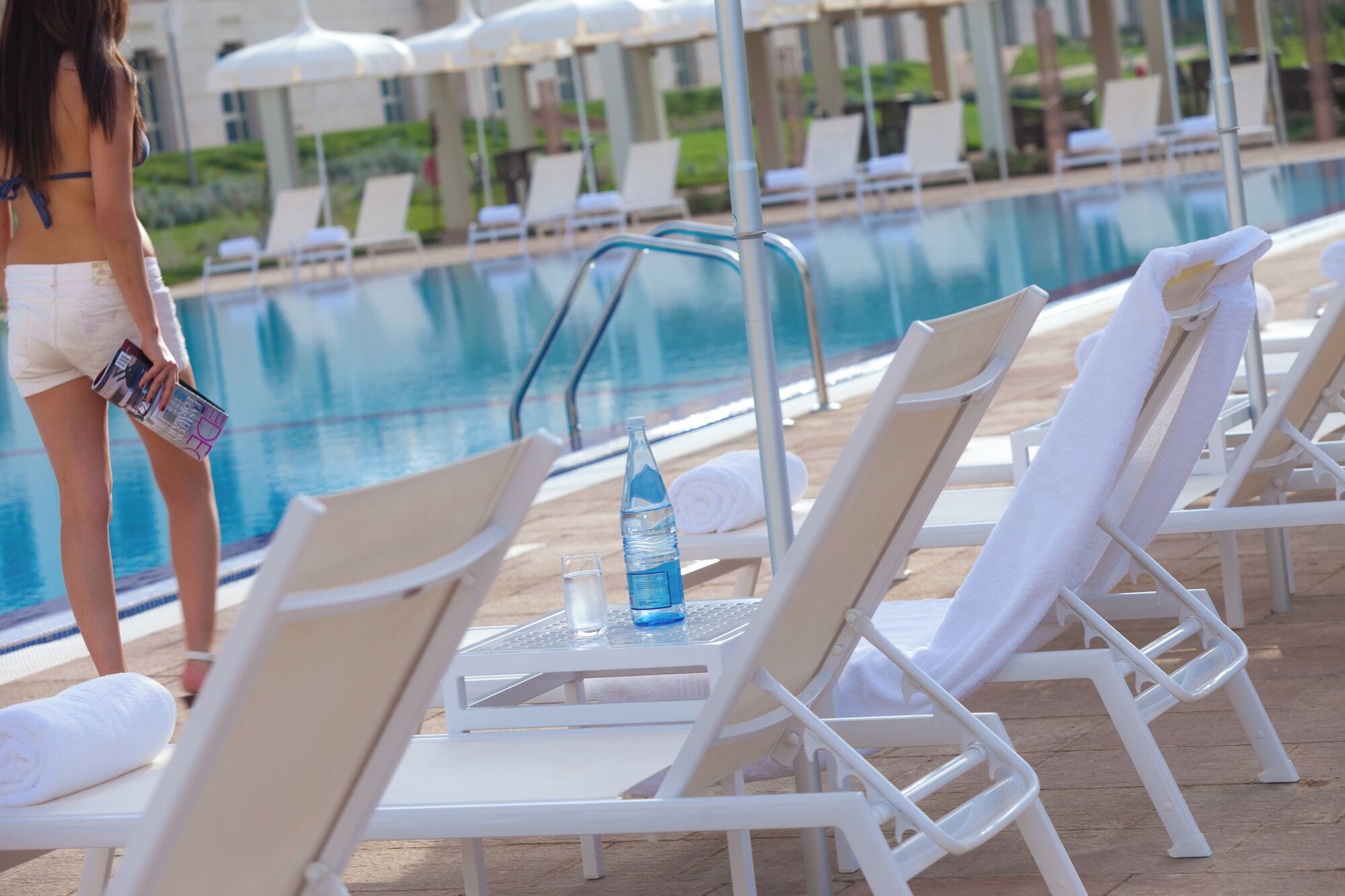 A poolside area with lounge chairs, a pool, and a person holding a magazine.