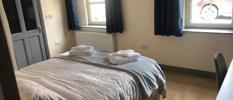 A hotel room with a bed, a desk, a chair, a radiator, and a window with curtains.