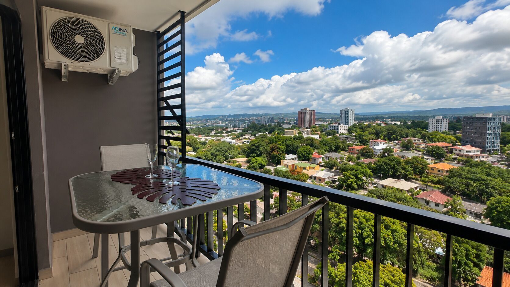 Un balcón con mesa y sillas, con vistas a un paisaje urbano con edificios y áreas verdes.
