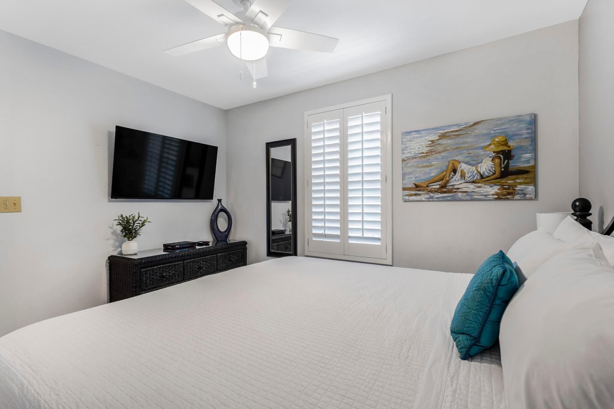 A bedroom with a large bed, a ceiling fan, a flat-screen TV, a painting, and a window with blinds.