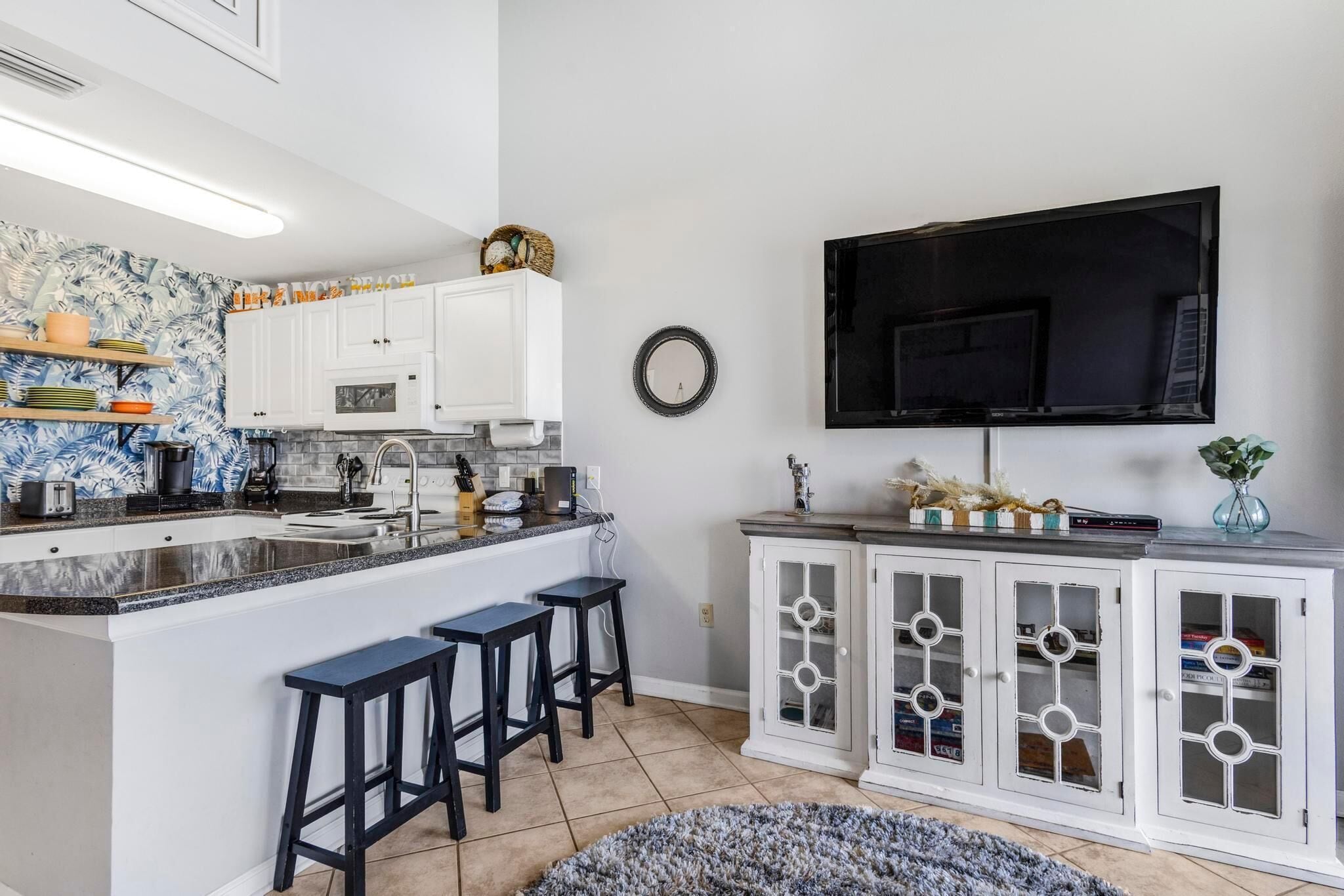 A modern kitchen with a central island, bar stools, a flat-screen TV, and a decorative mirror.
