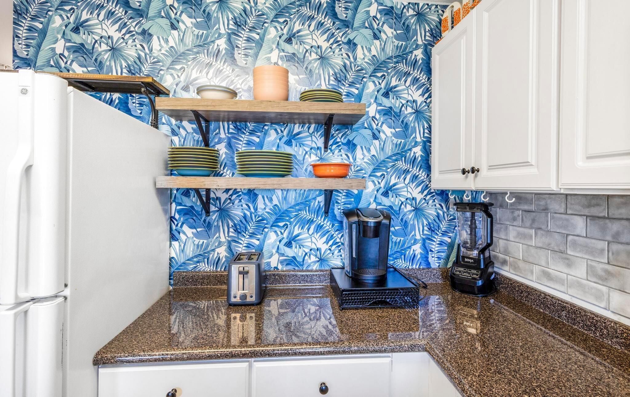 A kitchen with a tropical leaf wallpaper, a white refrigerator, a countertop with a toaster, a blender, and stacked plates on shelves.
