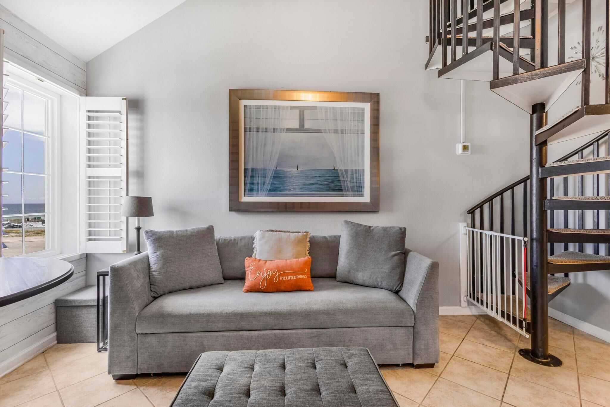 A living room with a grey sofa, a coffee table, a framed picture of a seascape, a staircase, and a window with blinds.