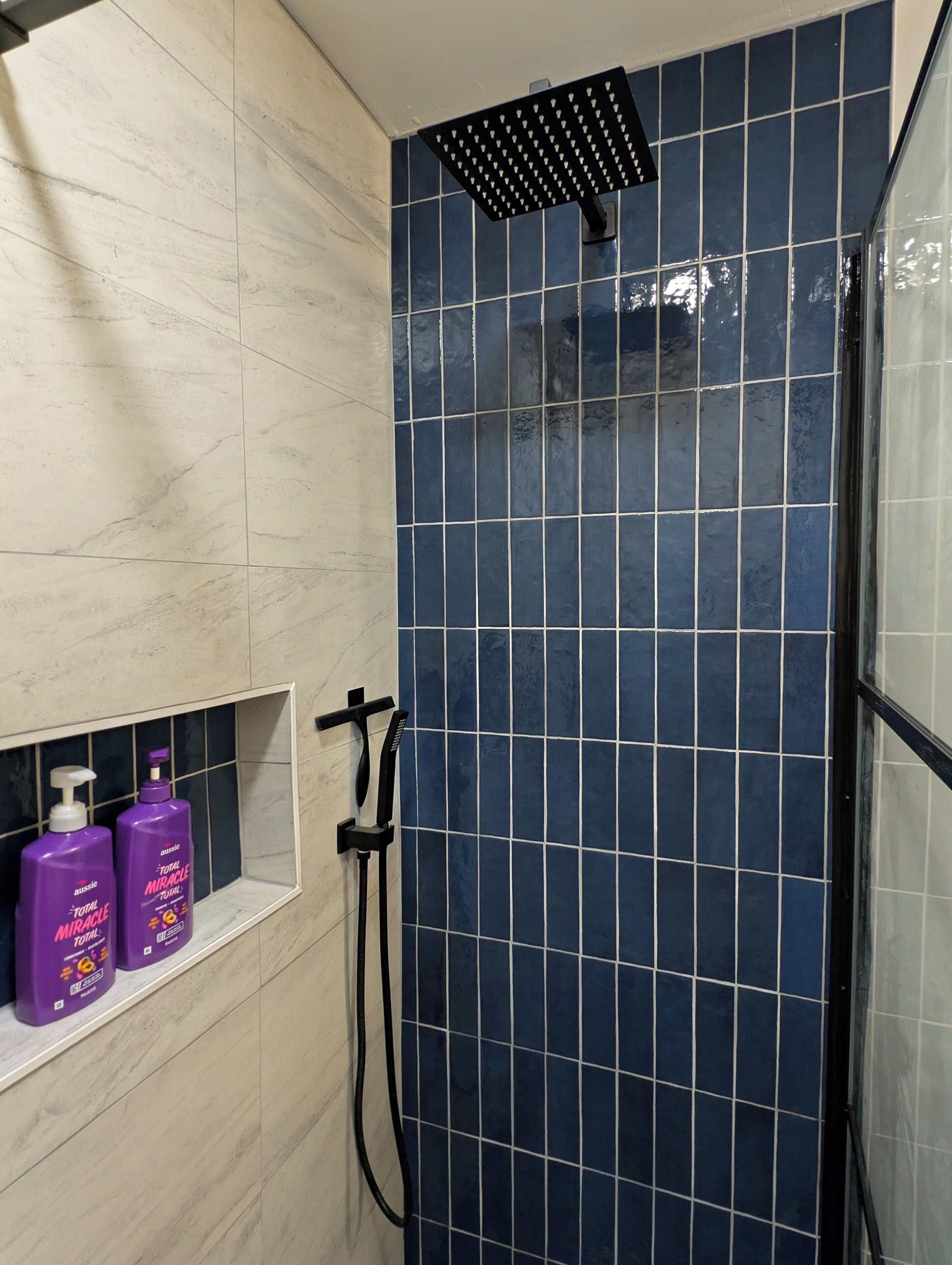 A bathroom with a shower area featuring blue tiles, a white marble wall, and a shelf with two bottles of Total Miracle Total.