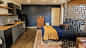 Quarto compacto com cozinha americana, cama com colcha estampada, ventilador de teto e divisória decorativa.
