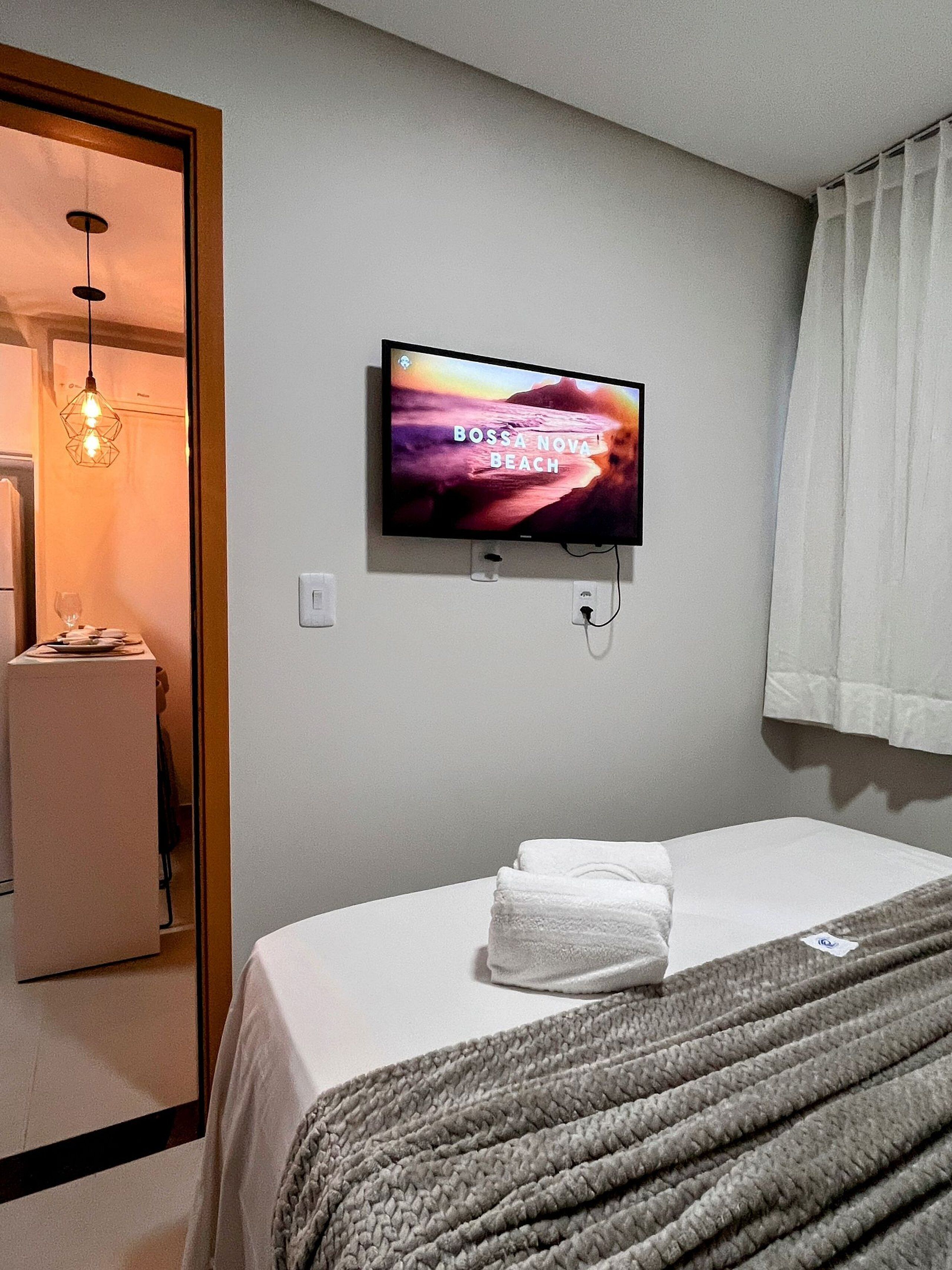 Une chambre d’hôtel avec un lit, un téléviseur fixé au mur, une serviette sur le lit et une fenêtre avec des rideaux.