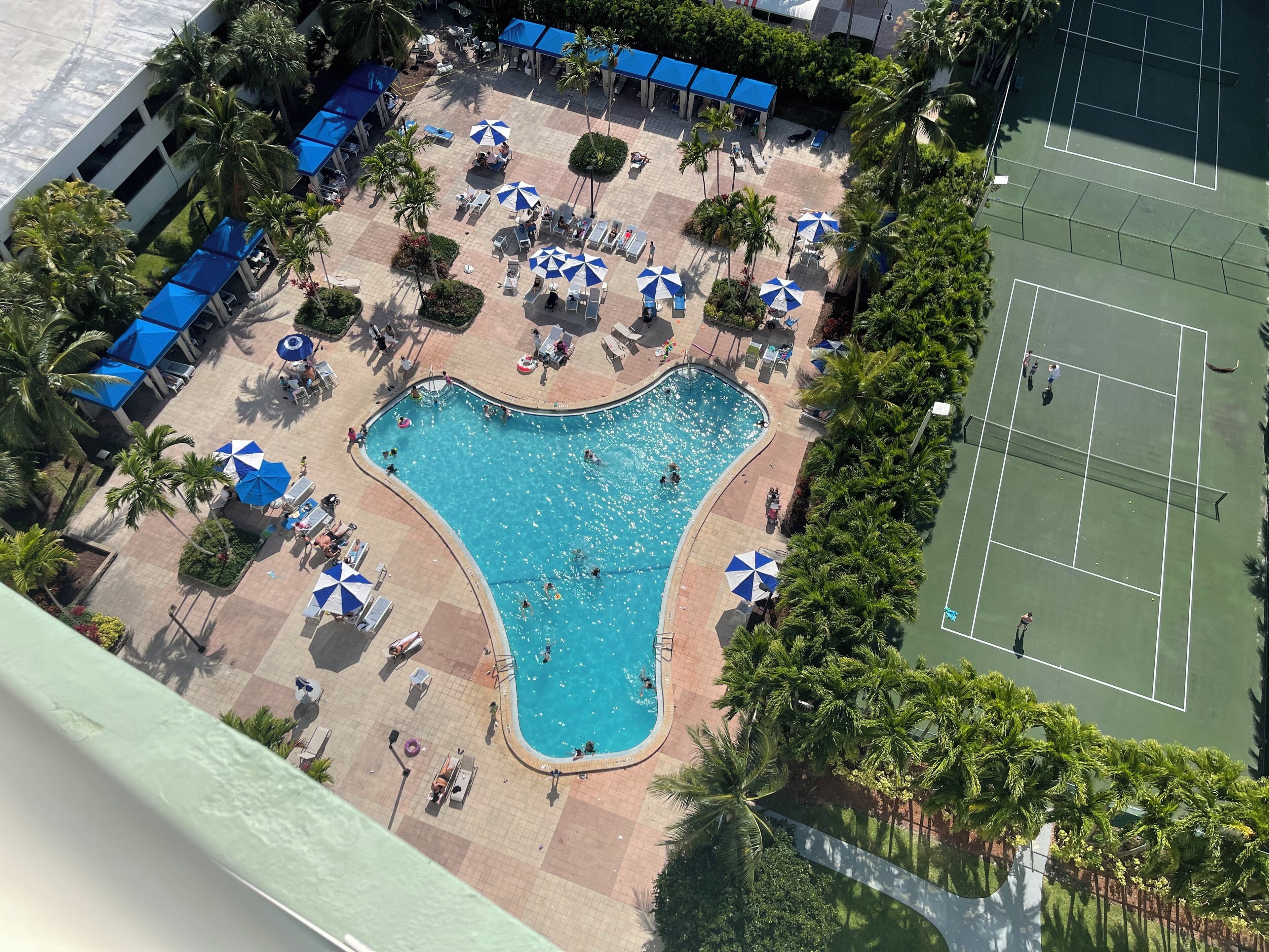 An aerial view of a resort with a swimming pool, tennis courts, and lounge areas with umbrellas.
