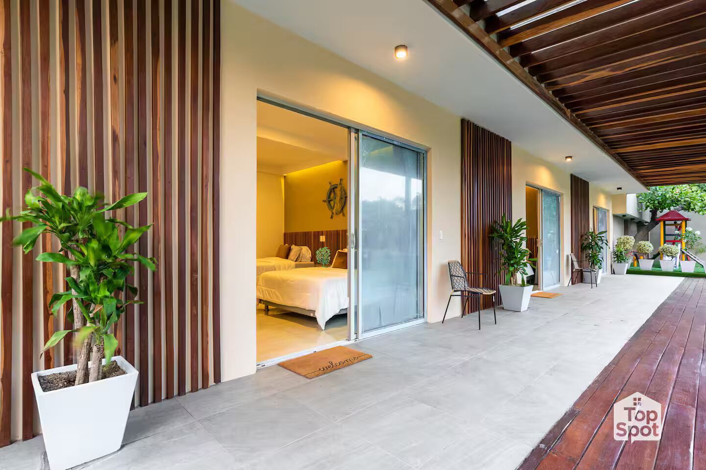 A modern hotel exterior with a covered outdoor area, potted plants, and a visible bedroom with a bed and window.