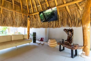 Um quarto com telhado de palha, um sofá, uma televisão montada no teto, uma escultura de madeira e uma cesta trançada.