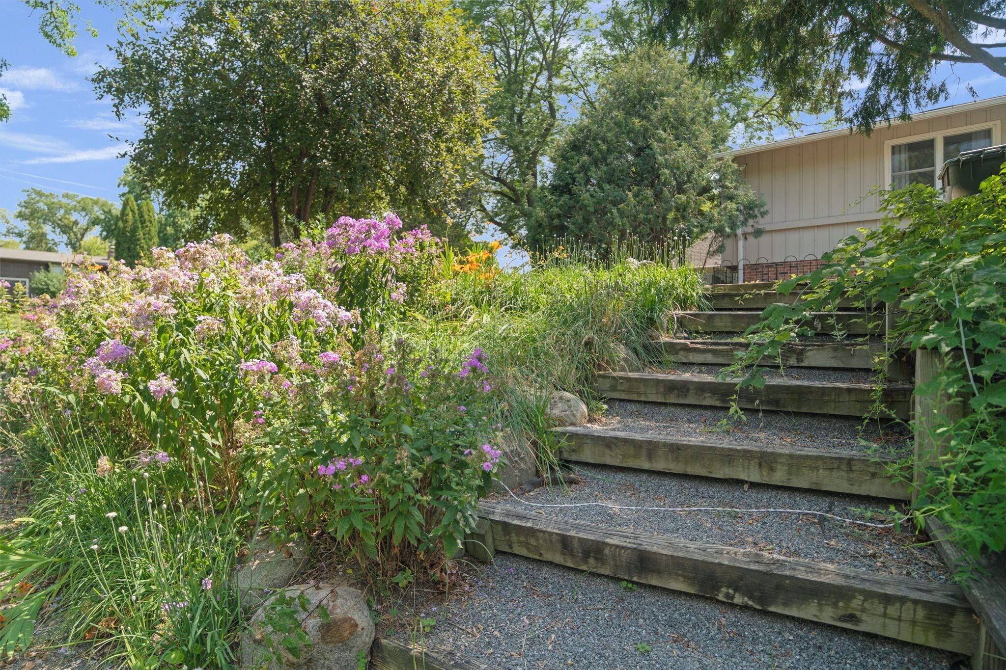 A set of wooden outdoor stairs leading up to a building, surrounded by lush greenery and flowering plants.