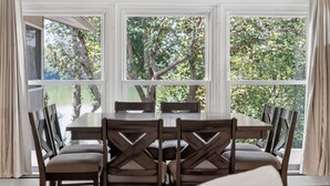 A dining area with a large table and chairs, large windows with curtains, and a view of trees. - Solara on the Lake (Plymouth)