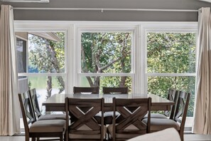 A dining area with a large table and chairs, large windows with curtains, and a view of trees. - Solara on the Lake (Plymouth)