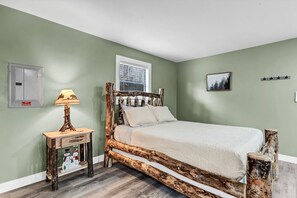 A bedroom with a rustic wooden bed frame, a bedside table with a lamp, a window with blinds, and a wall-mounted light switch.