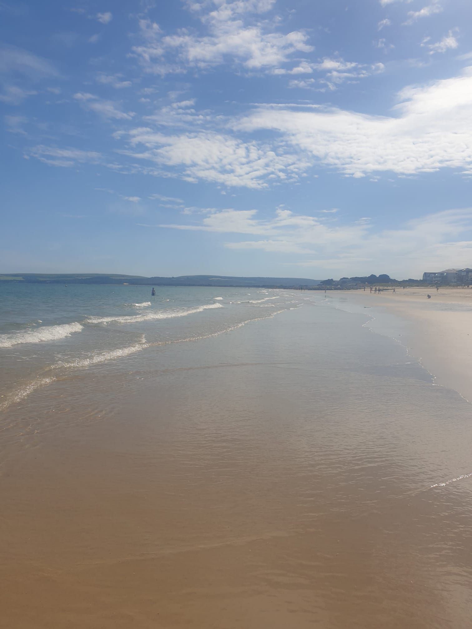 A wide sandy beach with gentle waves, a clear sky, and distant buildings.