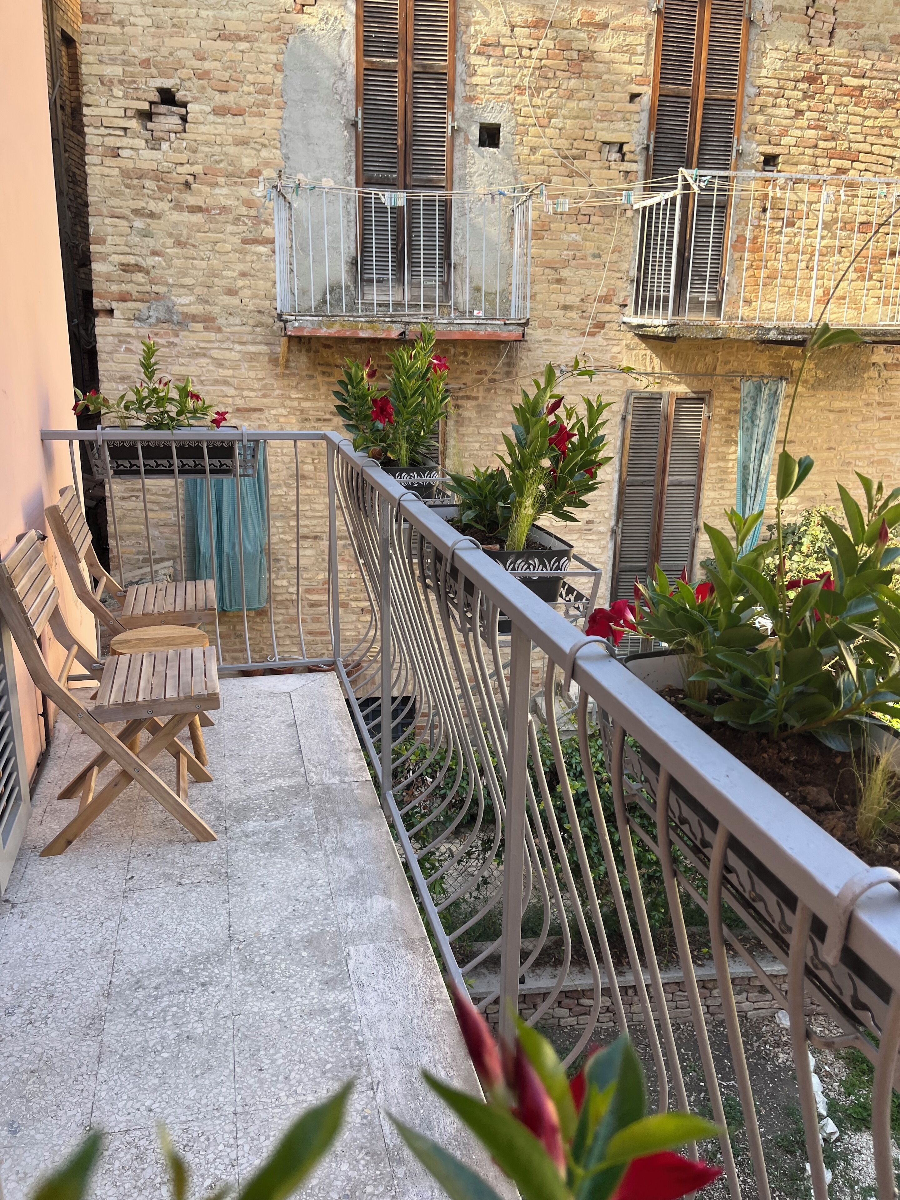 A balcony with a metal railing, wooden chairs, potted plants, and a stone wall.