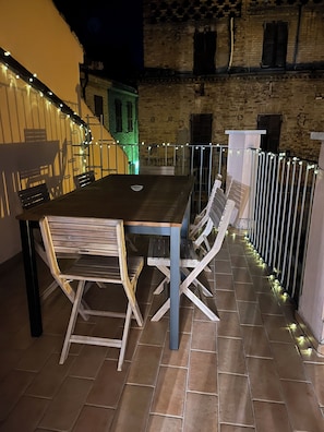 An outdoor patio with a wooden table and chairs, a metal railing, and a brick building in the background.