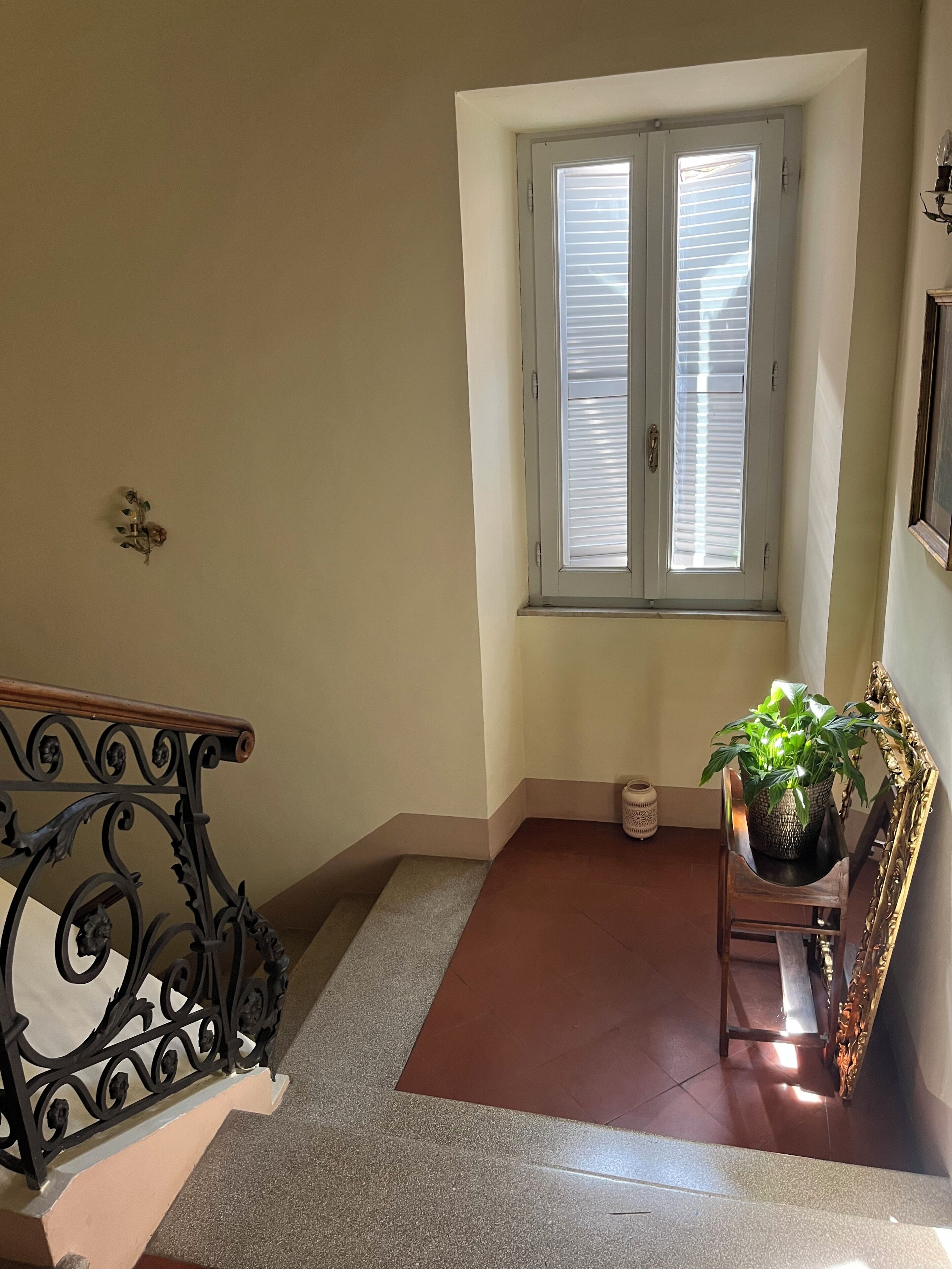 Una escalera con barandilla, una ventana con persianas, una planta en maceta y un espejo decorativo.