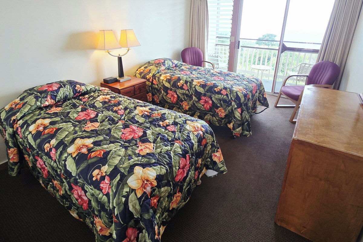 A hotel room with two beds, a desk, a chair, a ceiling fan, and a window with curtains.