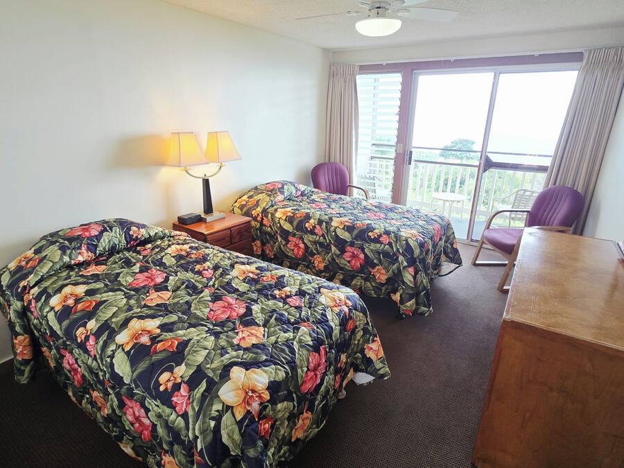 A hotel room with two beds, a desk, a chair, a ceiling fan, and a window with curtains.